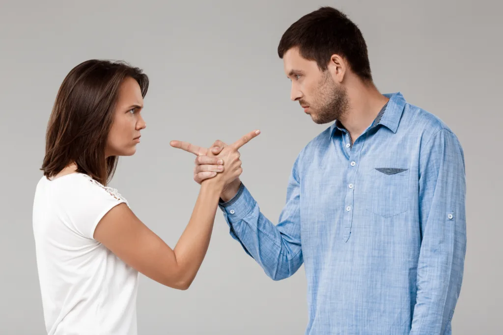 Young beautiful married couple posing, pointing and looking at each other with rancor over grey background.